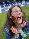 A woman with dramatic zombie makeup, including a torn cheek effect, screaming on a green soccer field with blue stadium seats in the background.